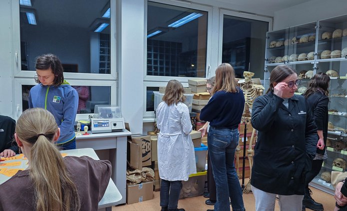 Grupa ludzi rozmawiająca w sali na tle okien i gablot oraz pudeł z materiałem kostnym